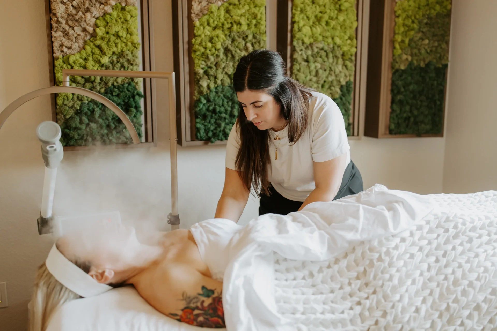 A beautician applies steam treatment to a client during a facial, with green wall art in the background and a cozy, serene atmosphere