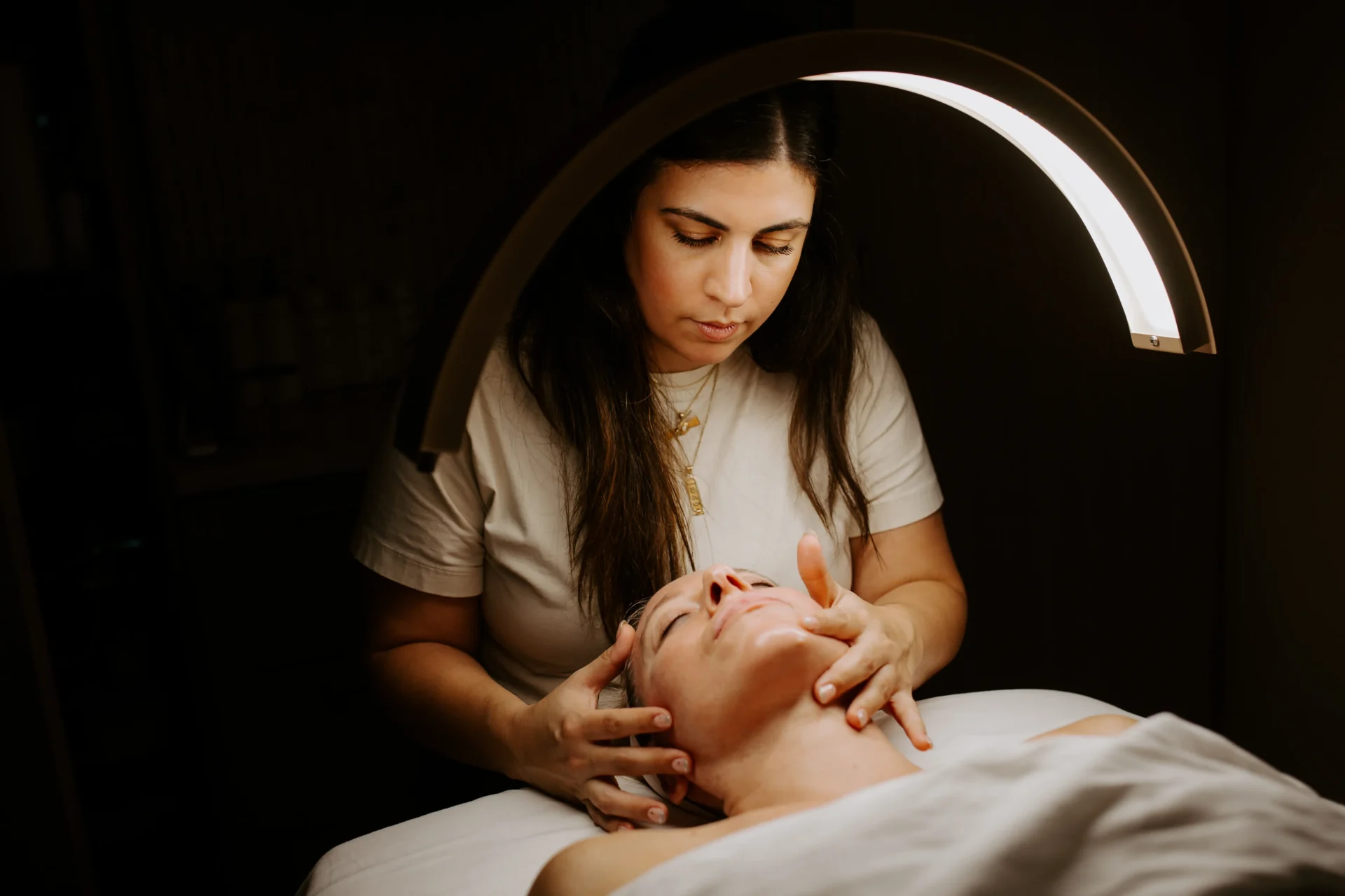 A person receives a facial treatment under a warm, arching lamp in a softly lit, relaxing environment