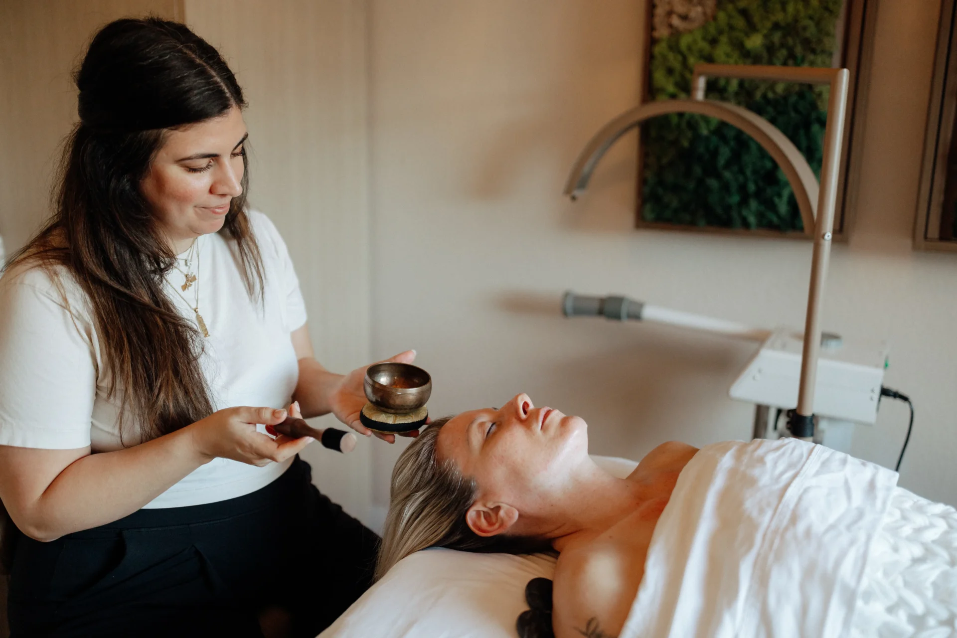 An aesthetician prepares to use a singing bowl near a client's head during a relaxing spa treatment