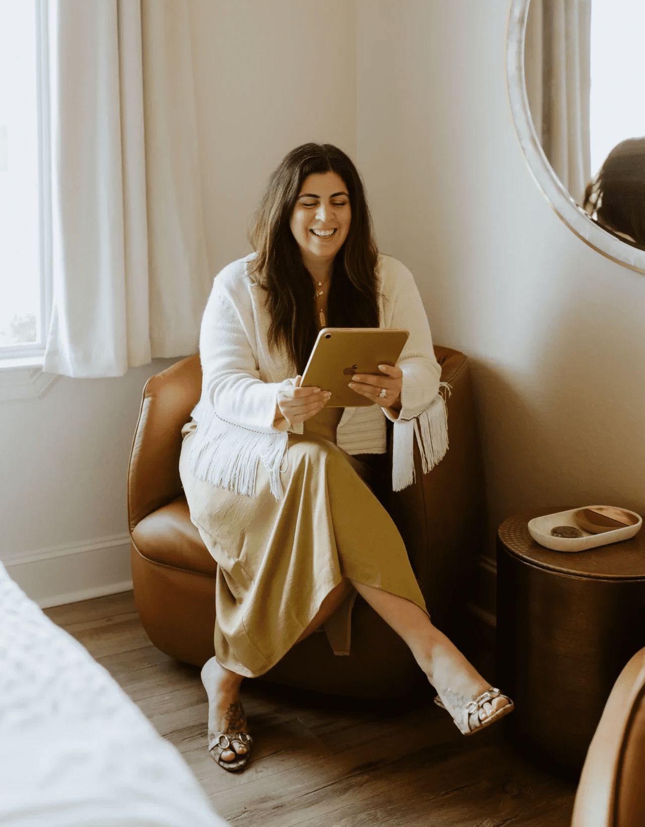 A smiling woman with long dark hair sits in a leather armchair, wearing a white fringe cardigan and gold dress while looking at a tablet